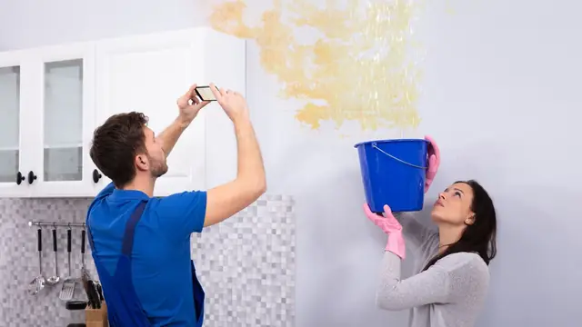 Man photographing a ceiling water stain while a woman holds a bucket